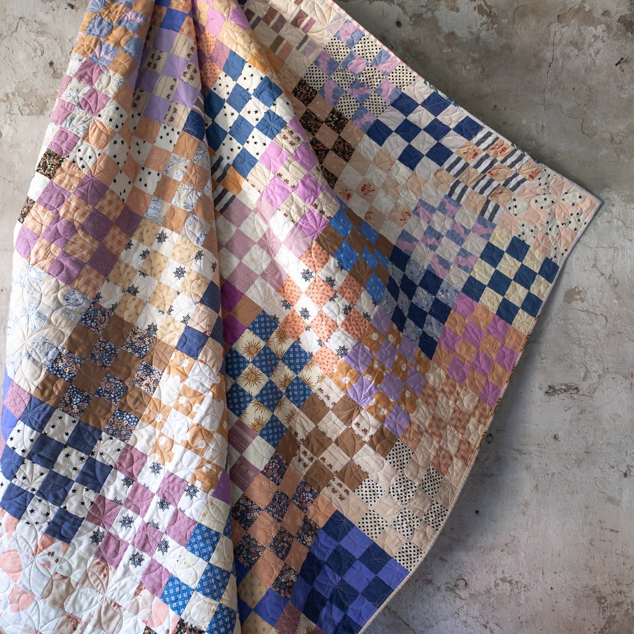 Scrappy 16 Patch Quilt hanging in front of a concrete wall.  The quilt is made primarily of blues, purples, oranges, and creams.