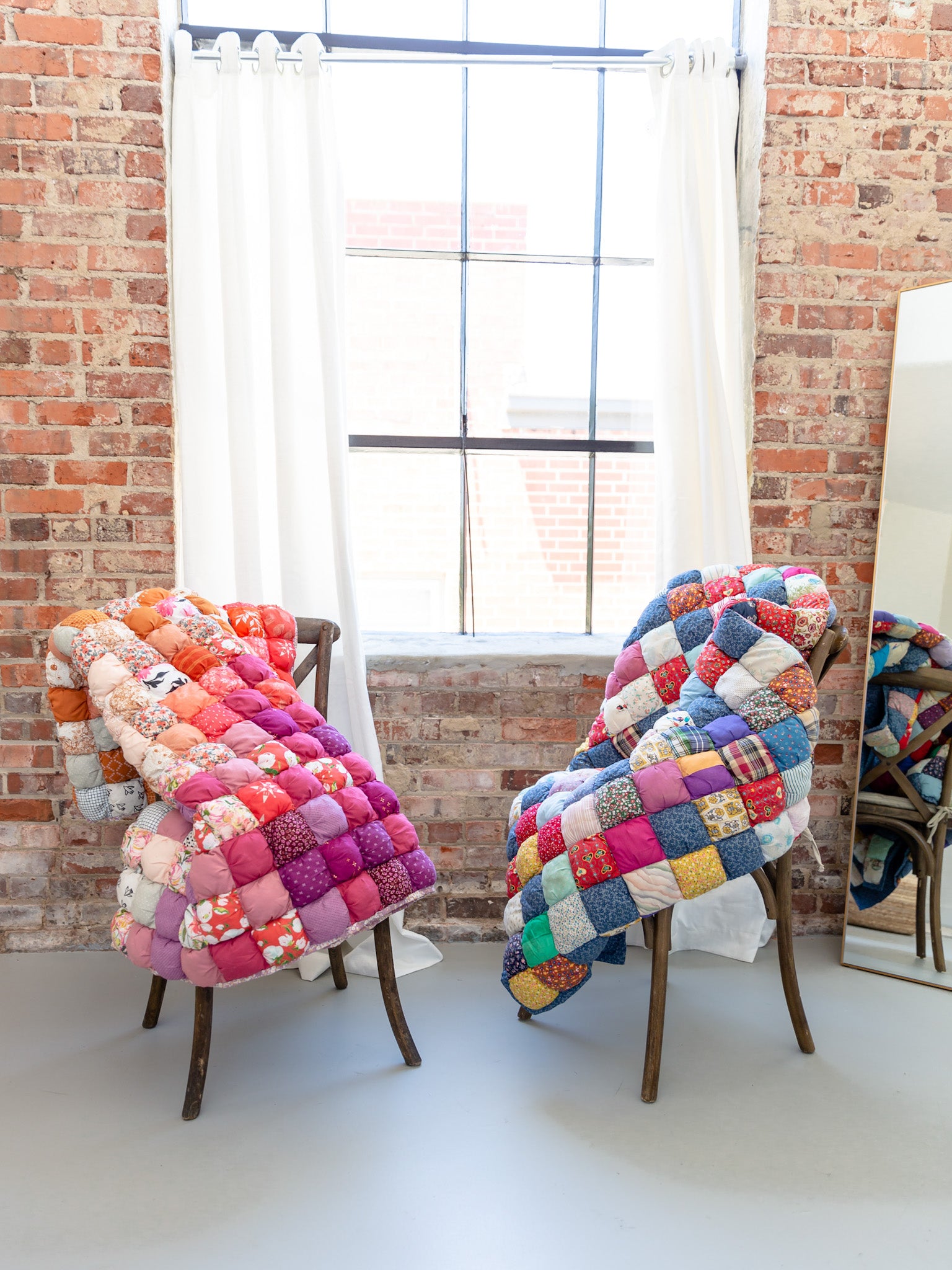 Photo of two puff quilts displayed on chairs by a window with abundant light streaming in.