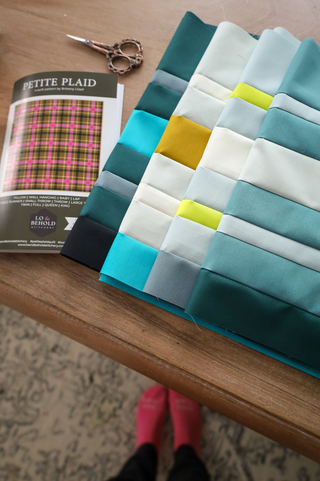Stacks of pieced fabric, scissors, and Petite Plaid quilt pattern by Lo & Behold Stitchery on a table. 
