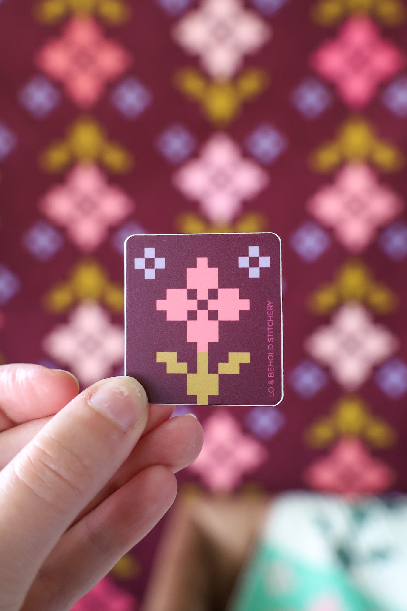 Hand holding a Pansy Patch Block Sticker by Lo & Behold Stitchery. Dark purple background with a medium pink pixel flower and two smaller purple grey block accent pixel flowers. 