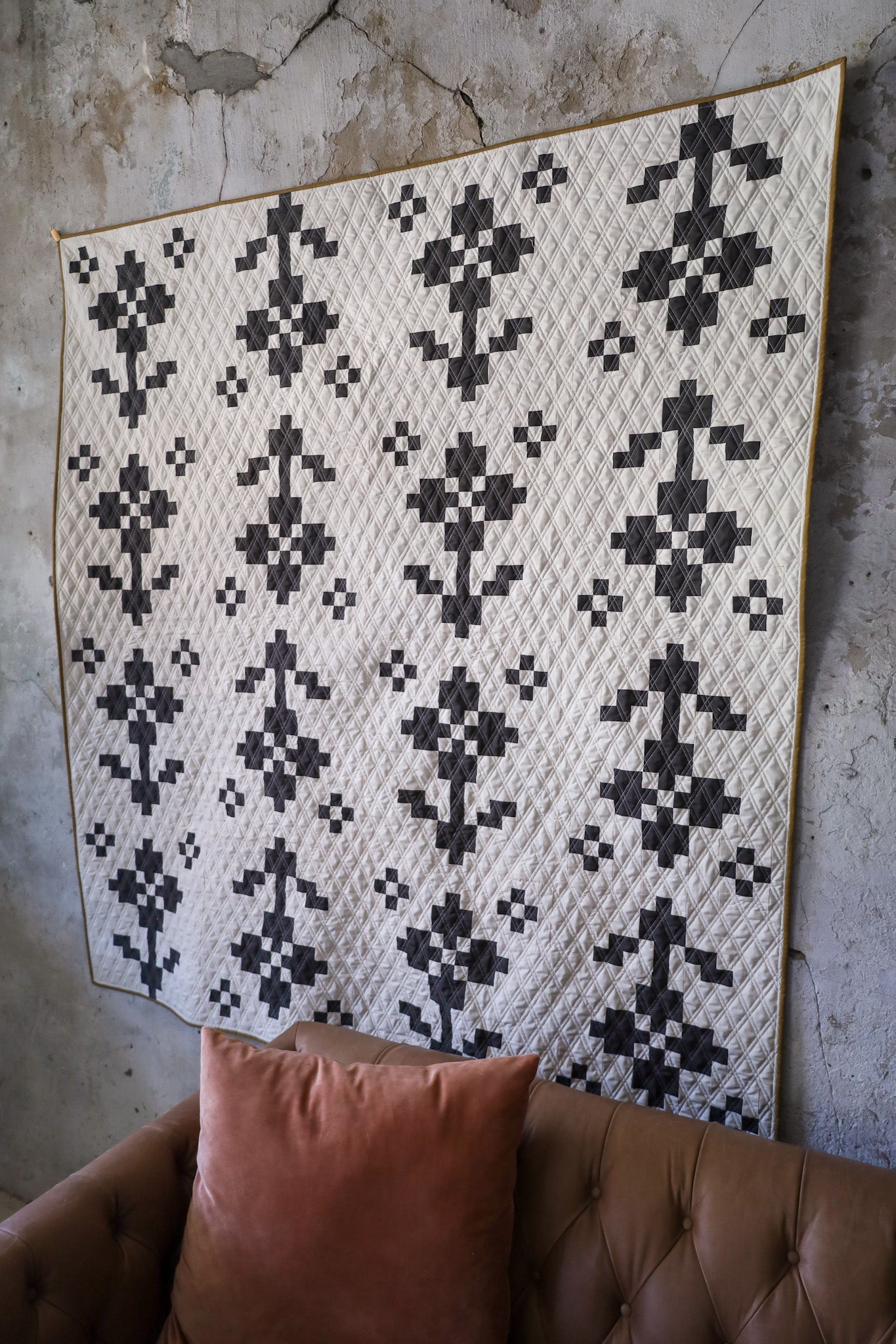 Pansy Patch Lava Rock sample quilt on a textured wall behind a brown couch and pink pillow